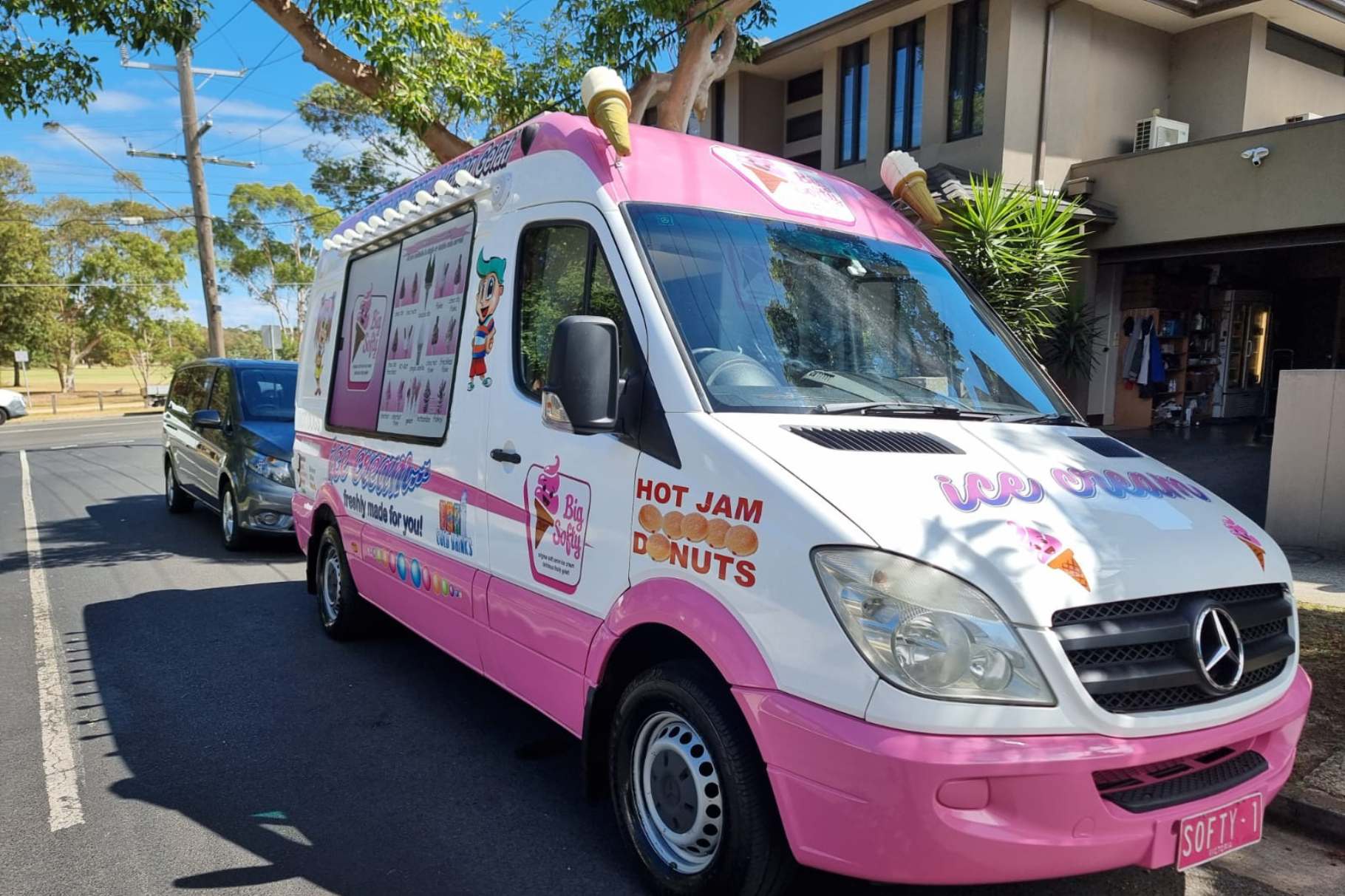 A pink and white ice cream van parked on the street under a tree, featuring colorful decals advertising ice cream, hot jam donuts, and a playful mascot.
