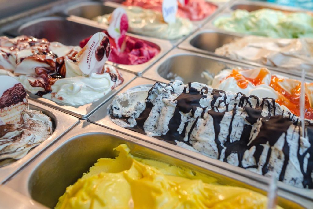 Assorted flavors of gelato in a display case with colorful toppings and signs.