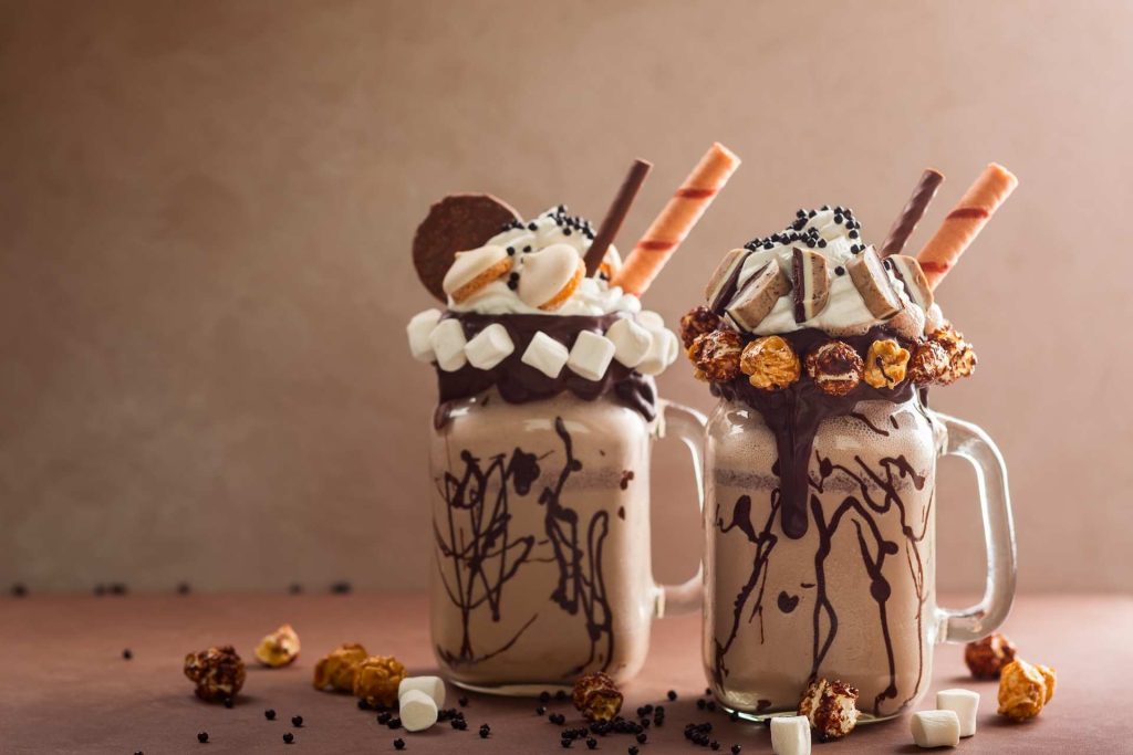 Two indulgent milkshakes in glass mugs, topped with whipped cream, chocolate drizzle, wafer sticks, marshmallows, cookies, and caramel popcorn.