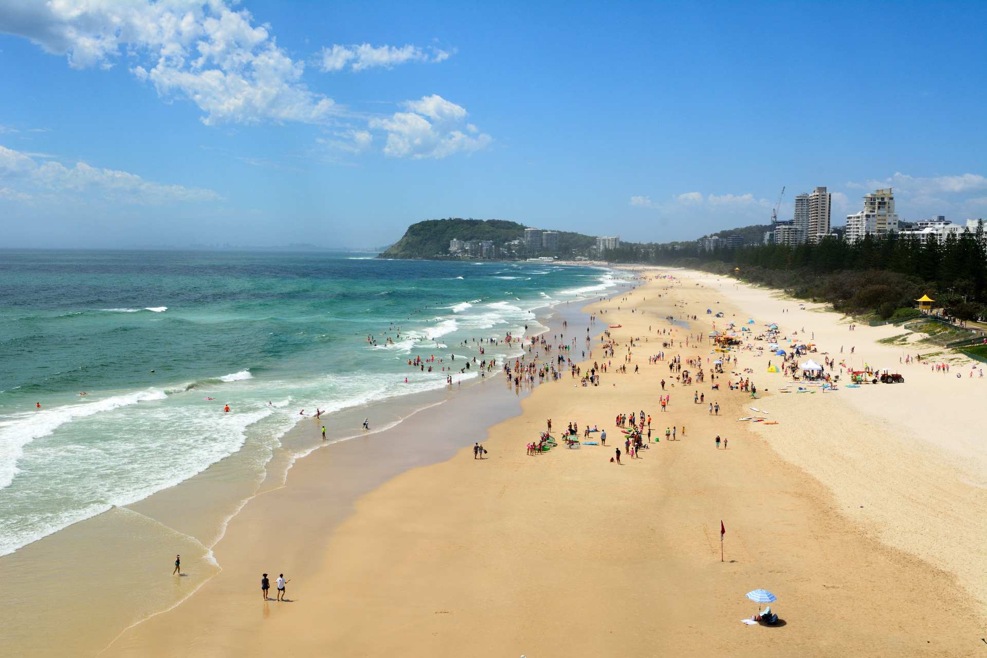 Aerial view of a crowded beach with people swimming and sunbathing, surrounded by clear blue skies and ocean waves. Tall buildings and trees in the background.