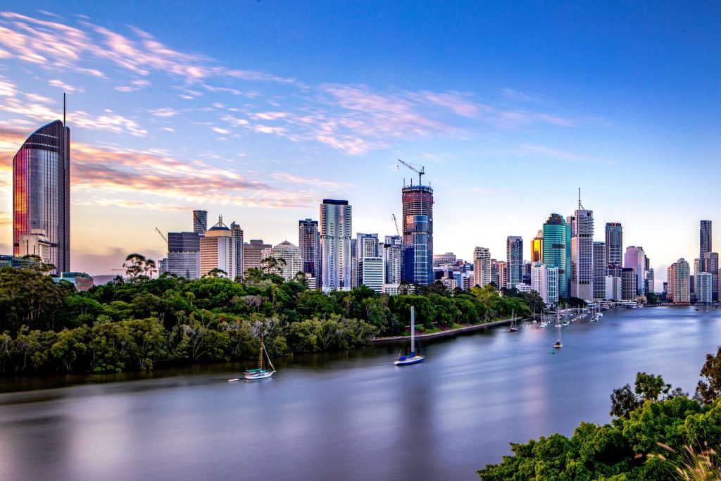 A panoramic view of a city skyline with tall buildings and construction cranes under a colorful evening sky, with a river in the foreground and lush greenery along the riverbank.