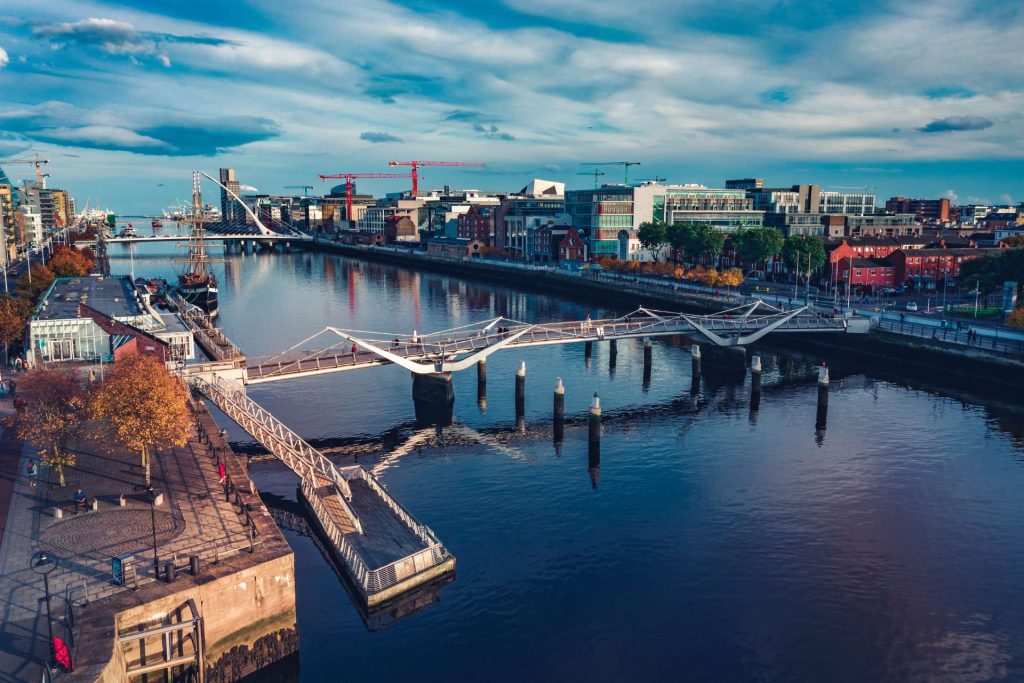 Aerial view of a river with bridges and cityscape, featuring modern buildings, cranes, and autumn trees under a blue sky.