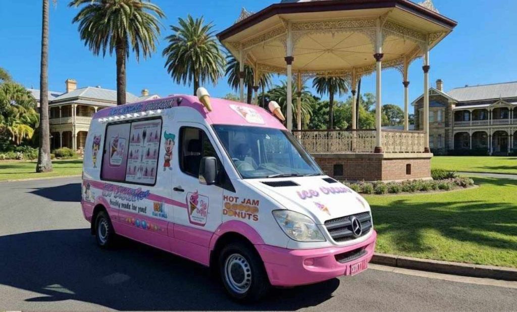 Pink and white ice cream van parked near a park gazebo with palm trees in the background.